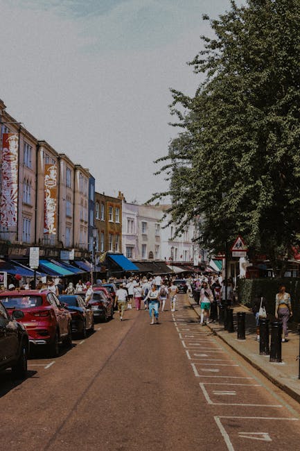 A narrow street in Putney, SW15, with parked cars lining both sides and pedestrians walking along the pavement. The buildings on the left feature colorful facades with retail shops and street-level aw
